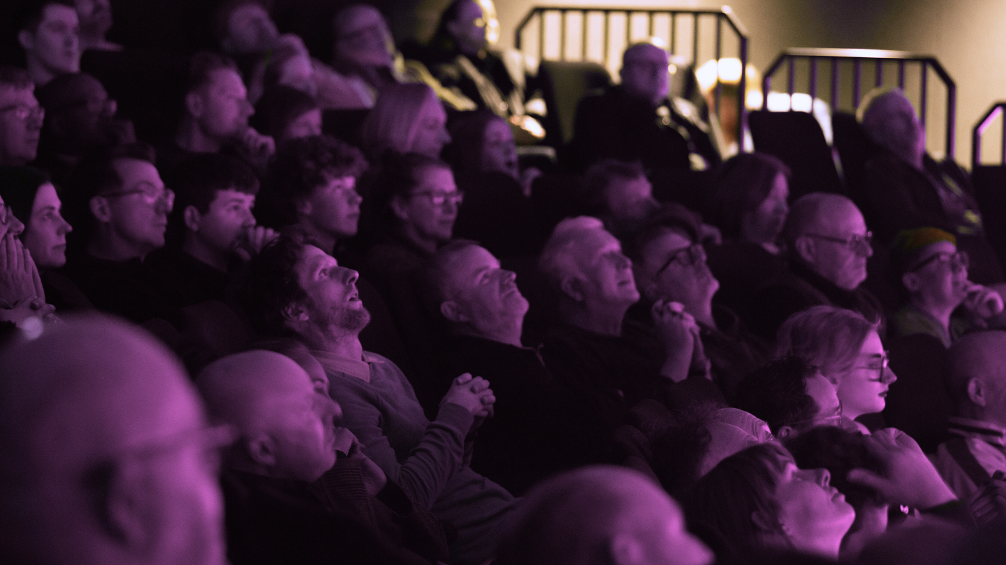 An audience watch a Space Dome Show with amazed expressions. The lighting is pink.