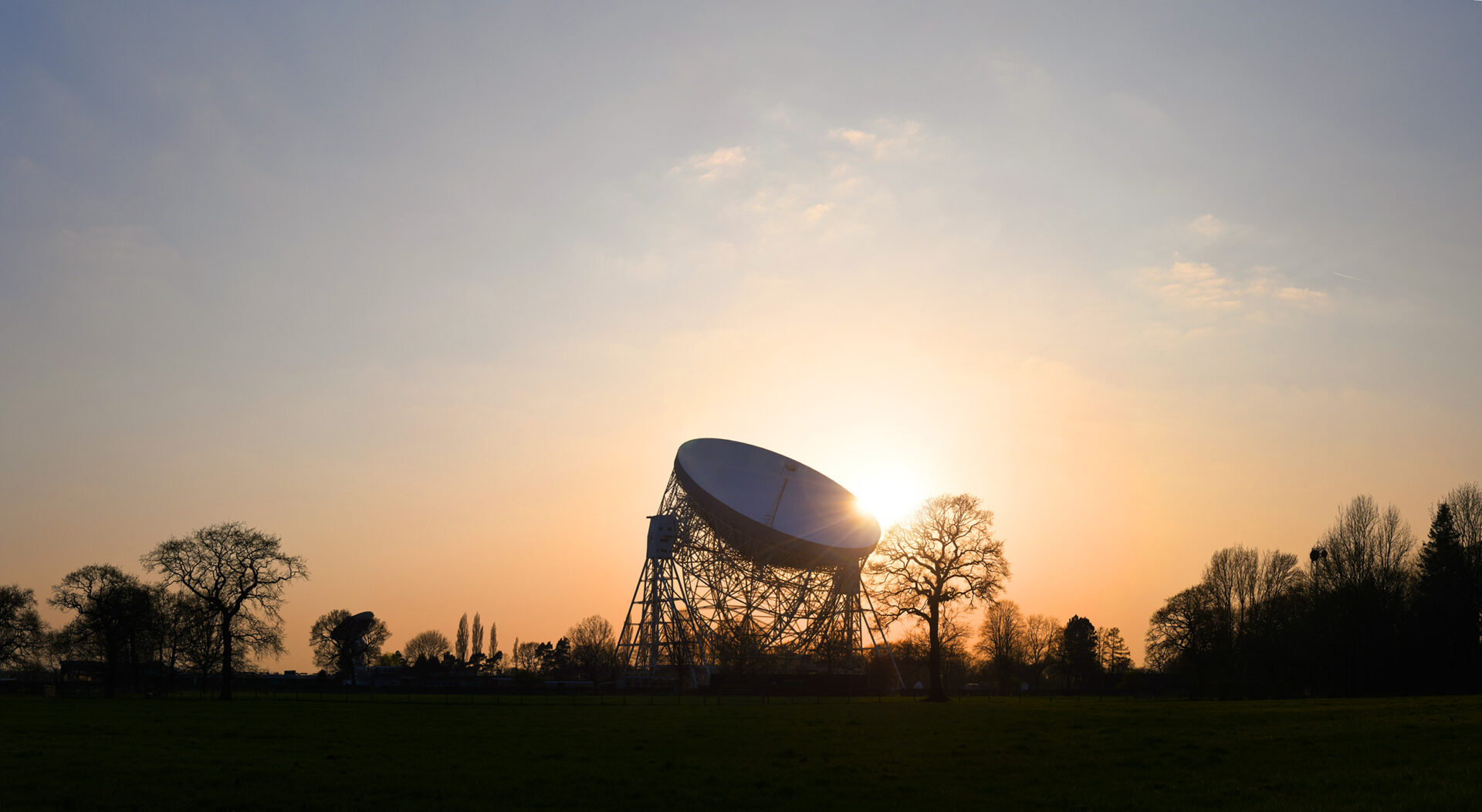 A photo of the Lovell Telescope at dusk, with the sun behind it.
