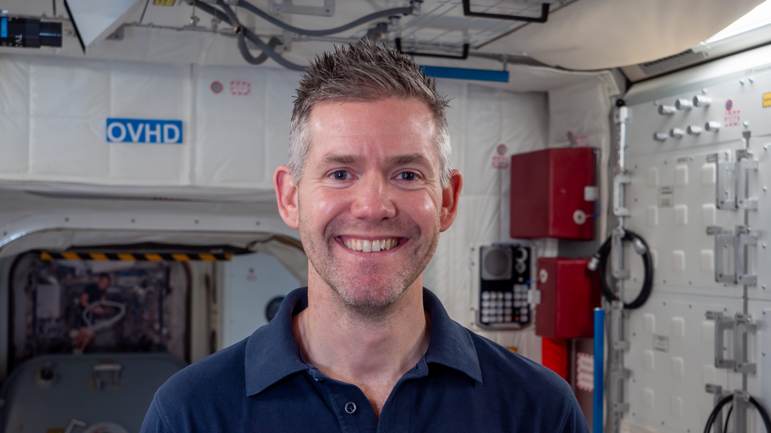 A photo of ESA Astronaut, John McFall, a smiling man with grey, spiked hair and a blue shirt.
