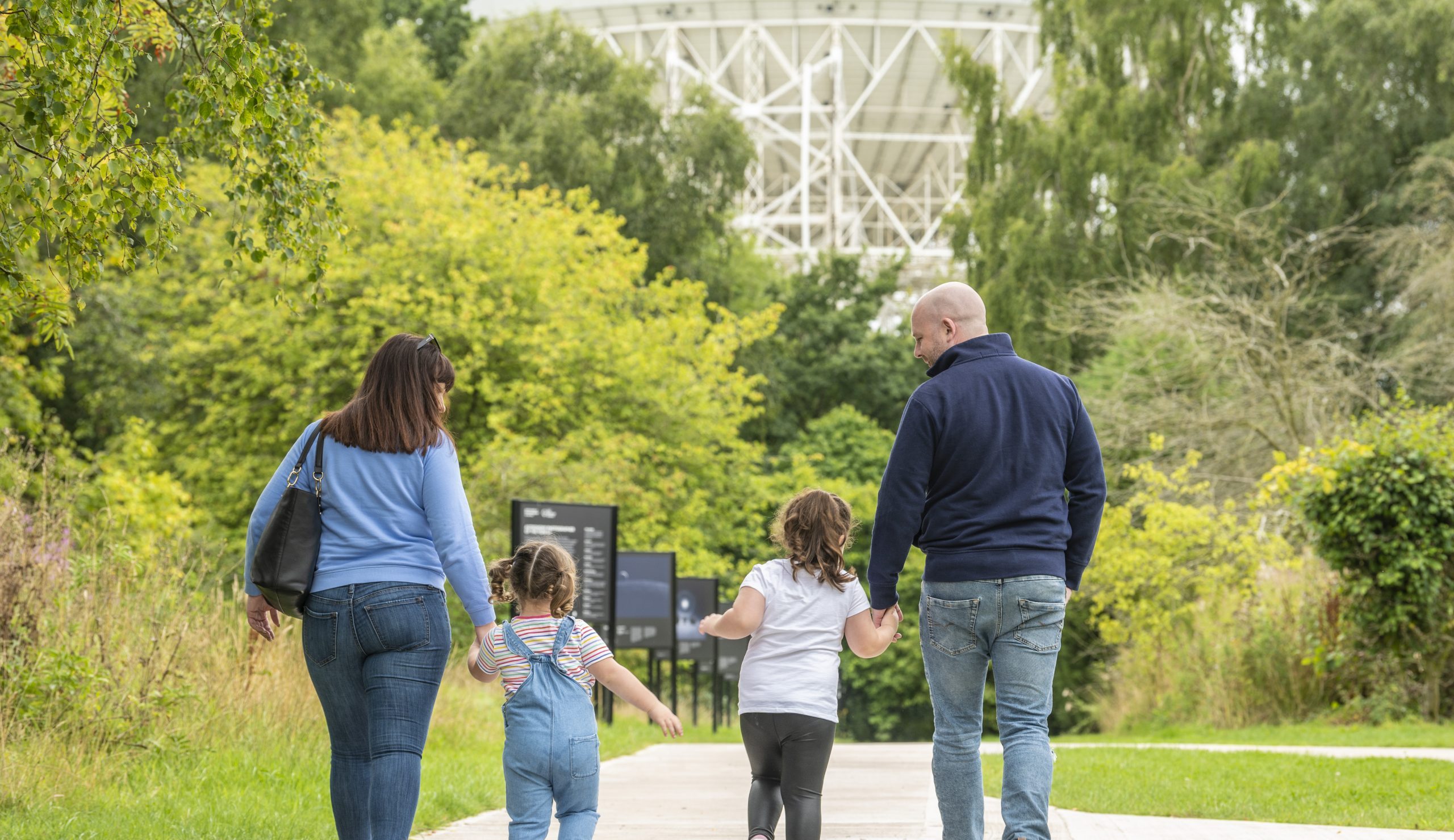 A photo of a family of four holding hands and walking towards the Lovell Telescope.
