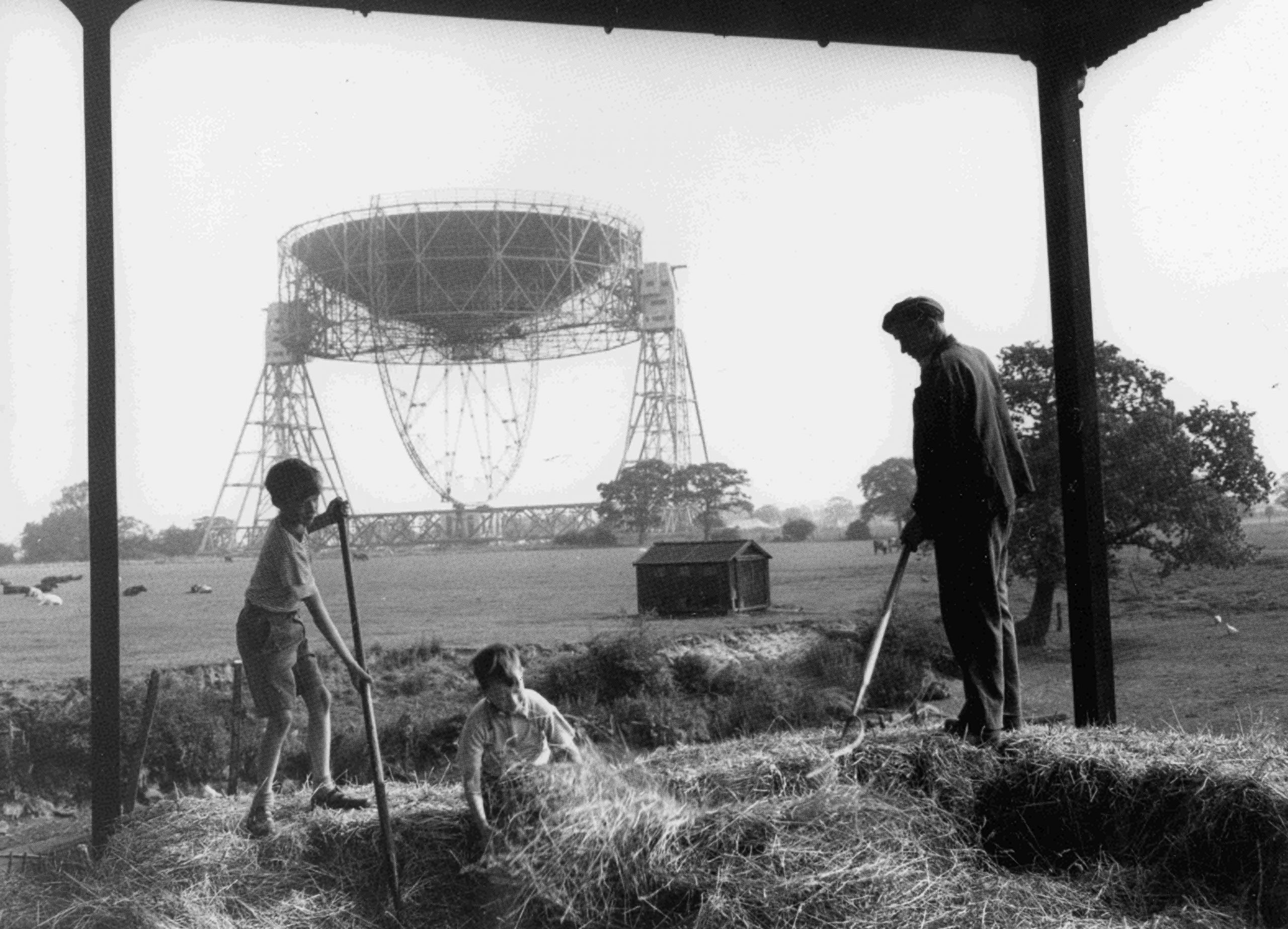 Black and white photograph of a man and two boys turning hay with pitchforks, with the telescope in the background.