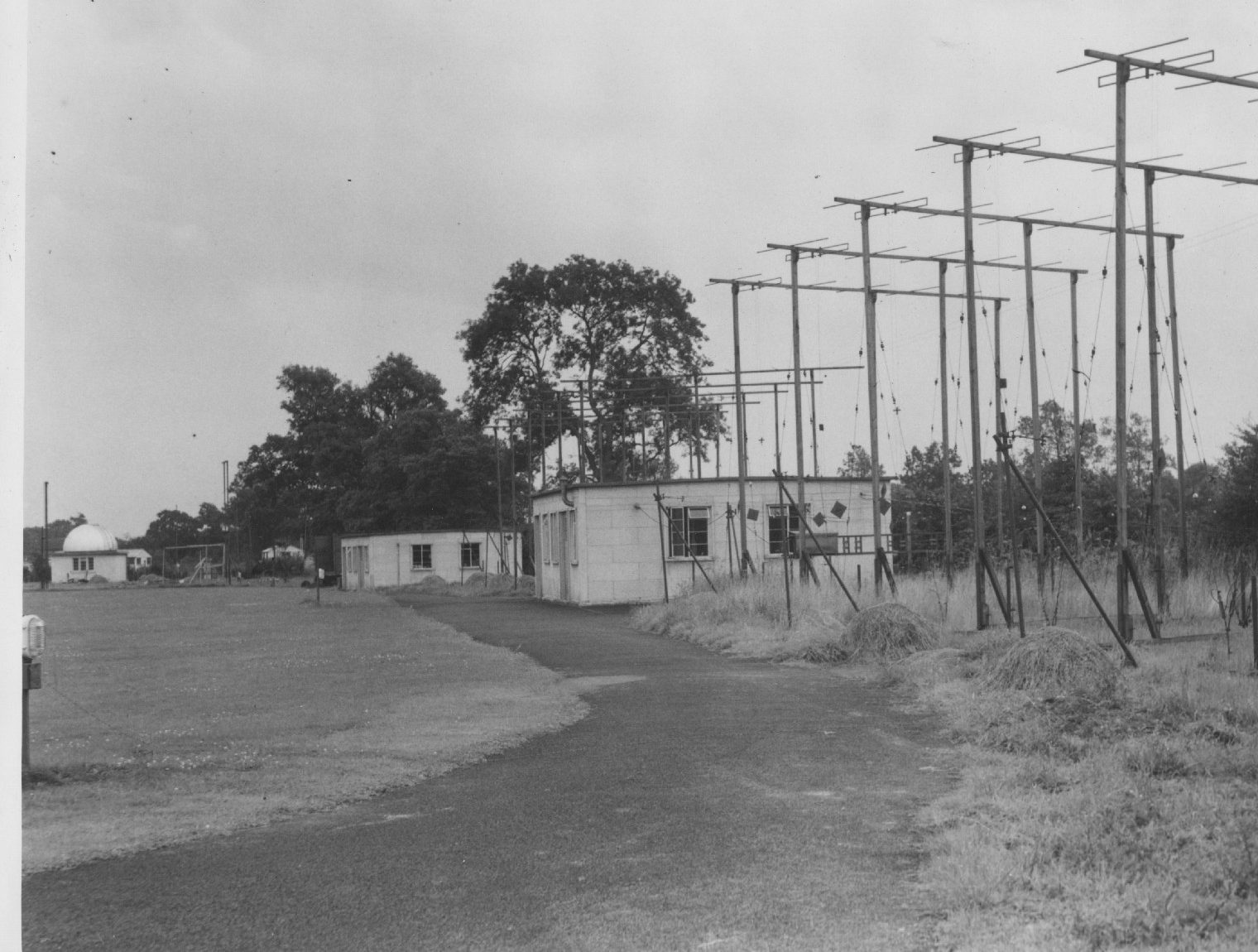 Black and white photograph of a view of the green with large yagi aerial array next to the Radiant hut, with Moon hut and optical observatory in the background.