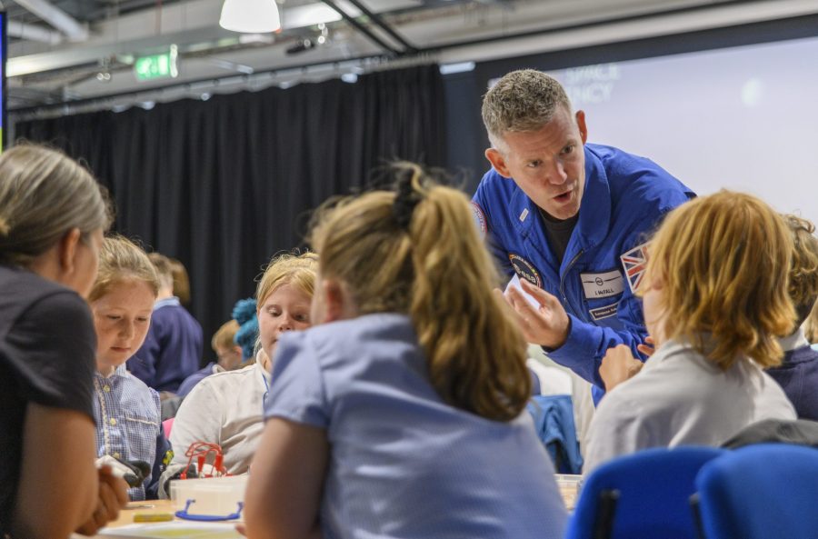 Astronaut John Mcfall, in a blue ESA spacesuit, talking to a table of primary school children.
