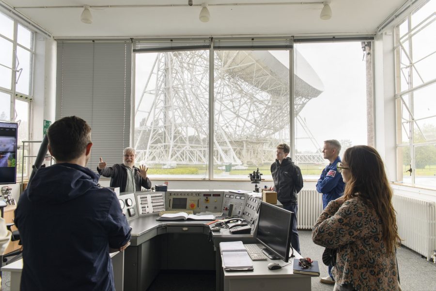 A group of people stood in the Jodrell Bank control room, the Lovell Telescope in the backdrop.