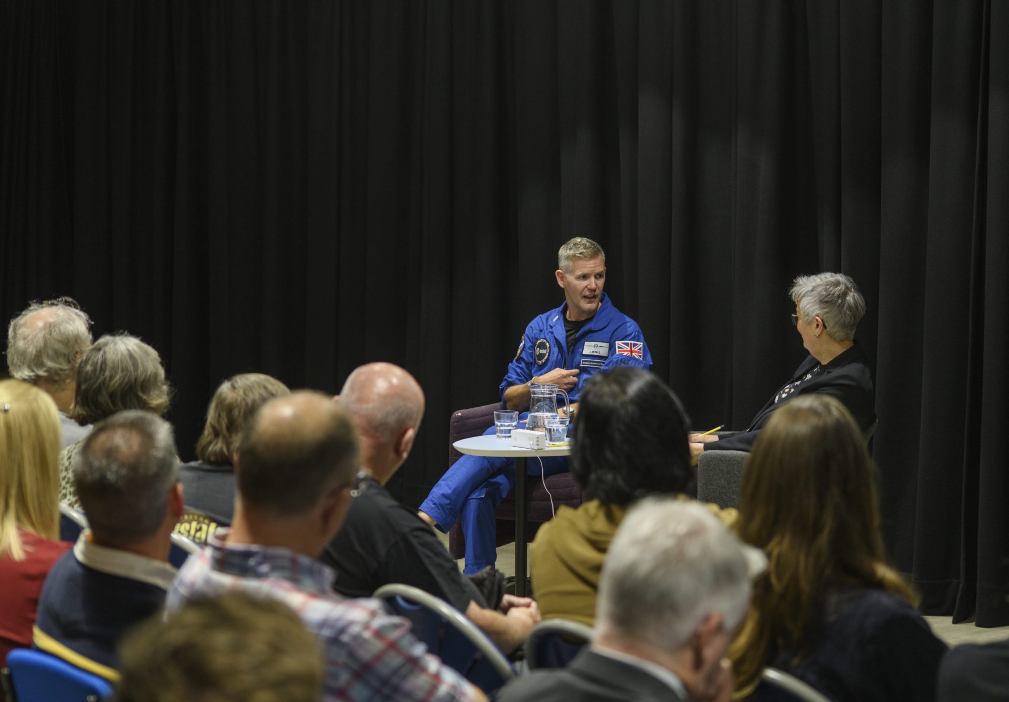 John McFall, a man in a blue ESA spacesuit, sat at a table in front of a crowd with JBCE Director, Teresa Anderson.