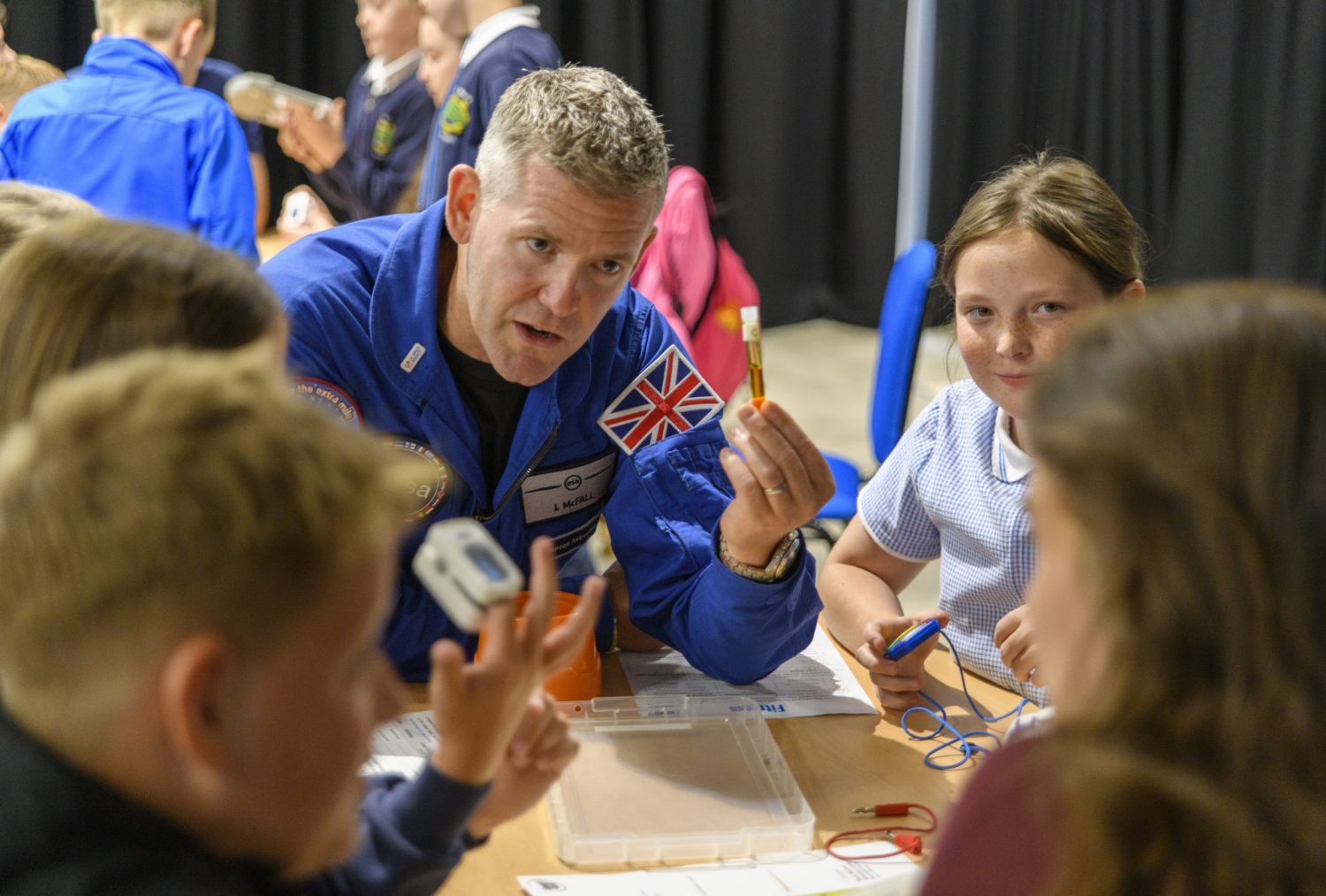 Astronaut John Mcfall, in a blue ESA spacesuit, talking to a table of primary school children.