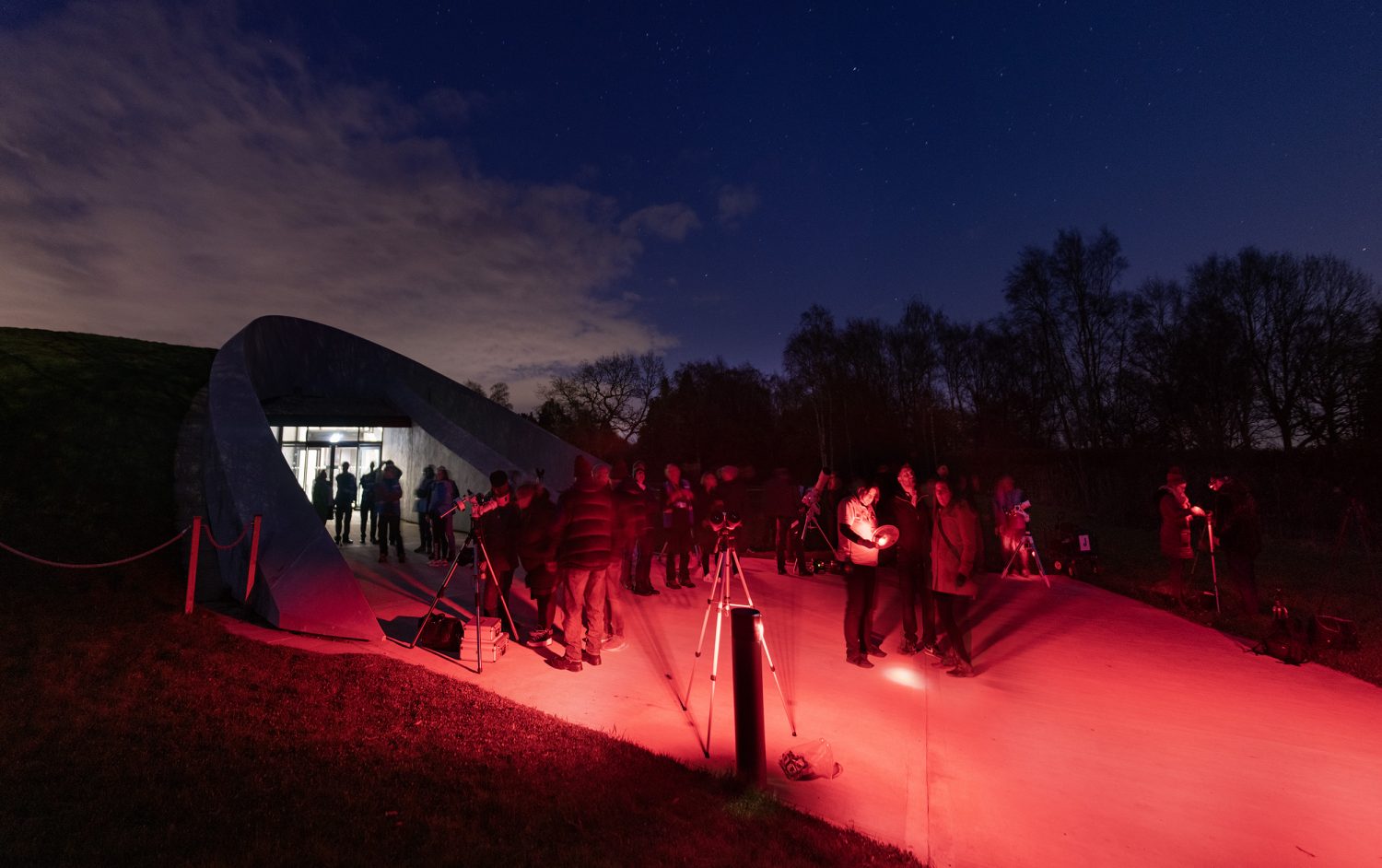 A photo of a group of people gathered at night in front of the First Light Pavilion. The photo is lit by red lighting.