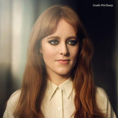 A headshot of Hannah Peel, a woman with red hair, a white shirt, and black eyeliner.