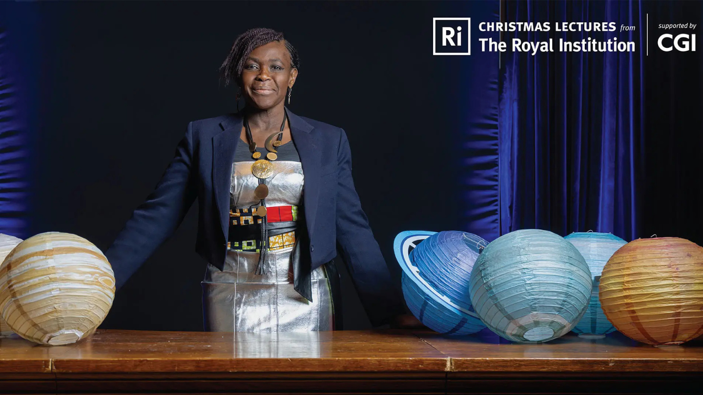 Dr Dame Maggie Aderin-Pocock stands behind a table of planets.