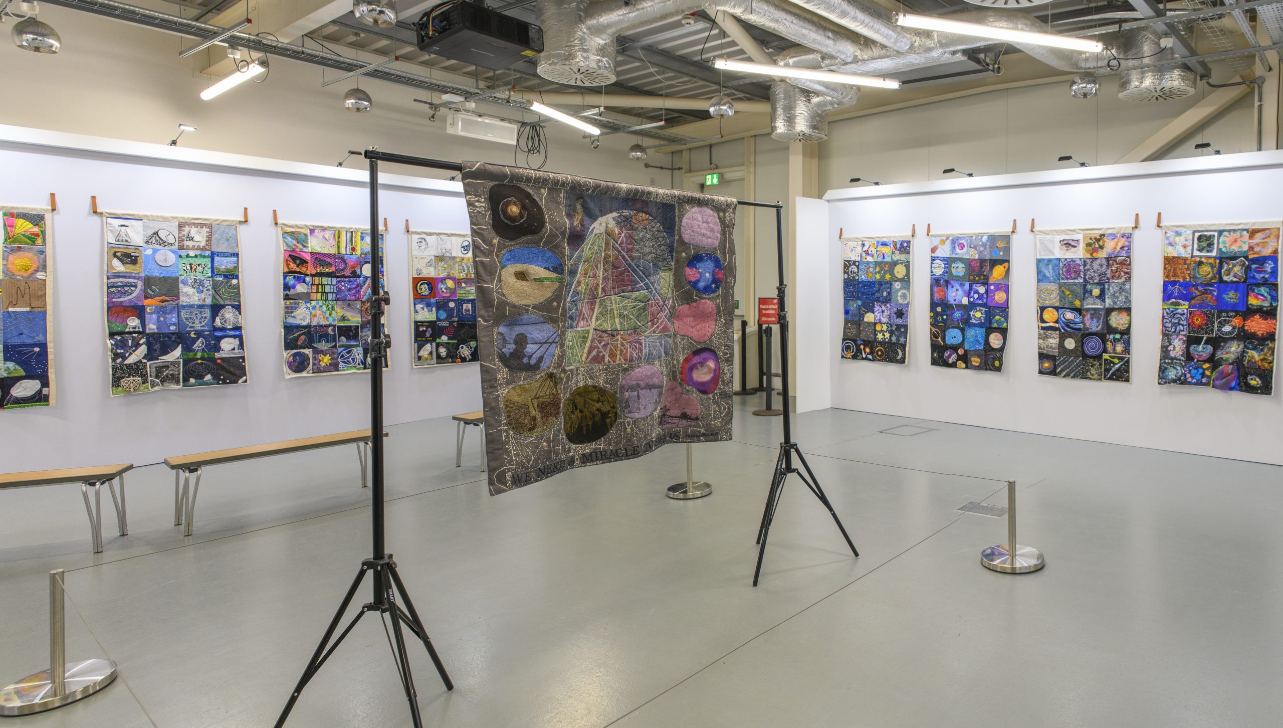 A room-wide photograph of the Cosmic Threads exhibition, including several hanging, textile quilts made up of public depictions of space and the Jodrell site, with artist Anne Kelly's quilt as the centrepiece.