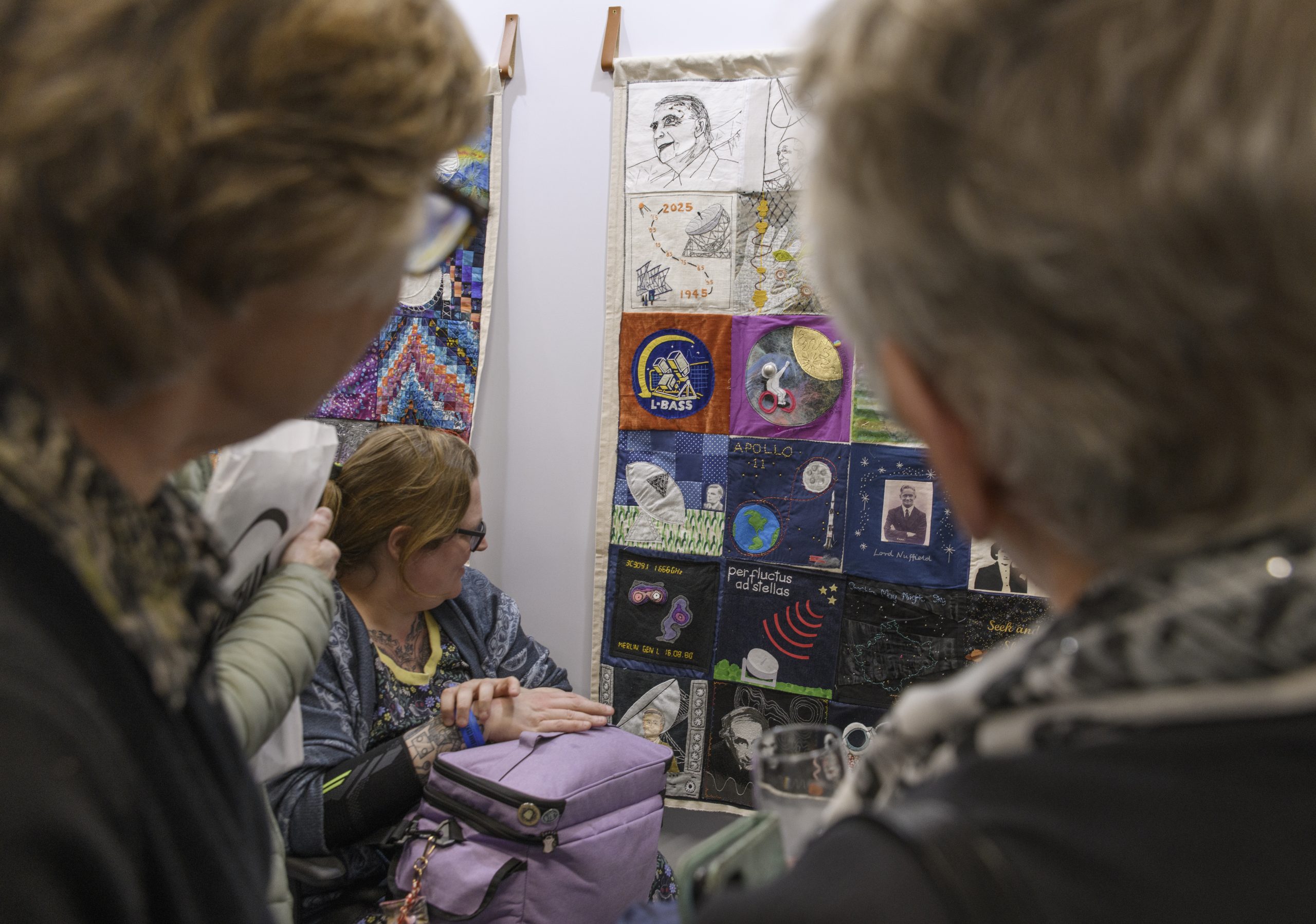 A group of visitors enjoy the colourful collection of hanging textile quilts, showing various squares depicting Jodrell Bank and space.