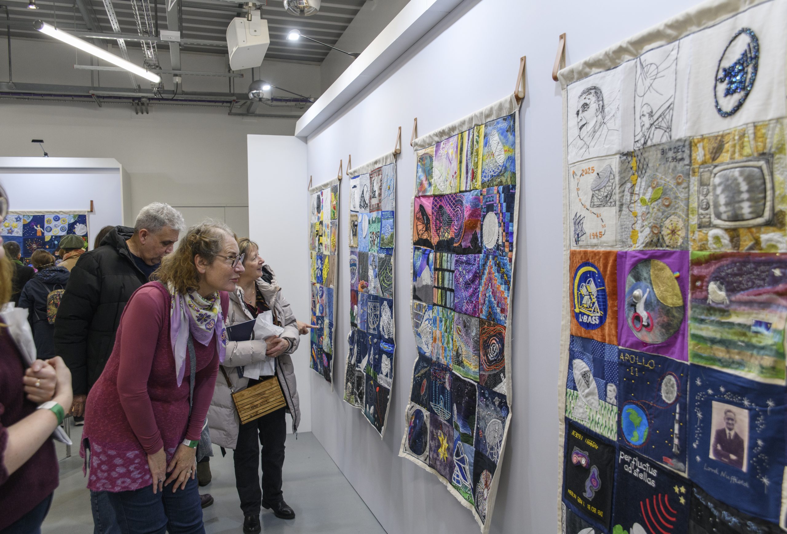 A group of visitors enjoy the colourful collection of hanging textile quilts, showing various squares depicting Jodrell Bank and space.