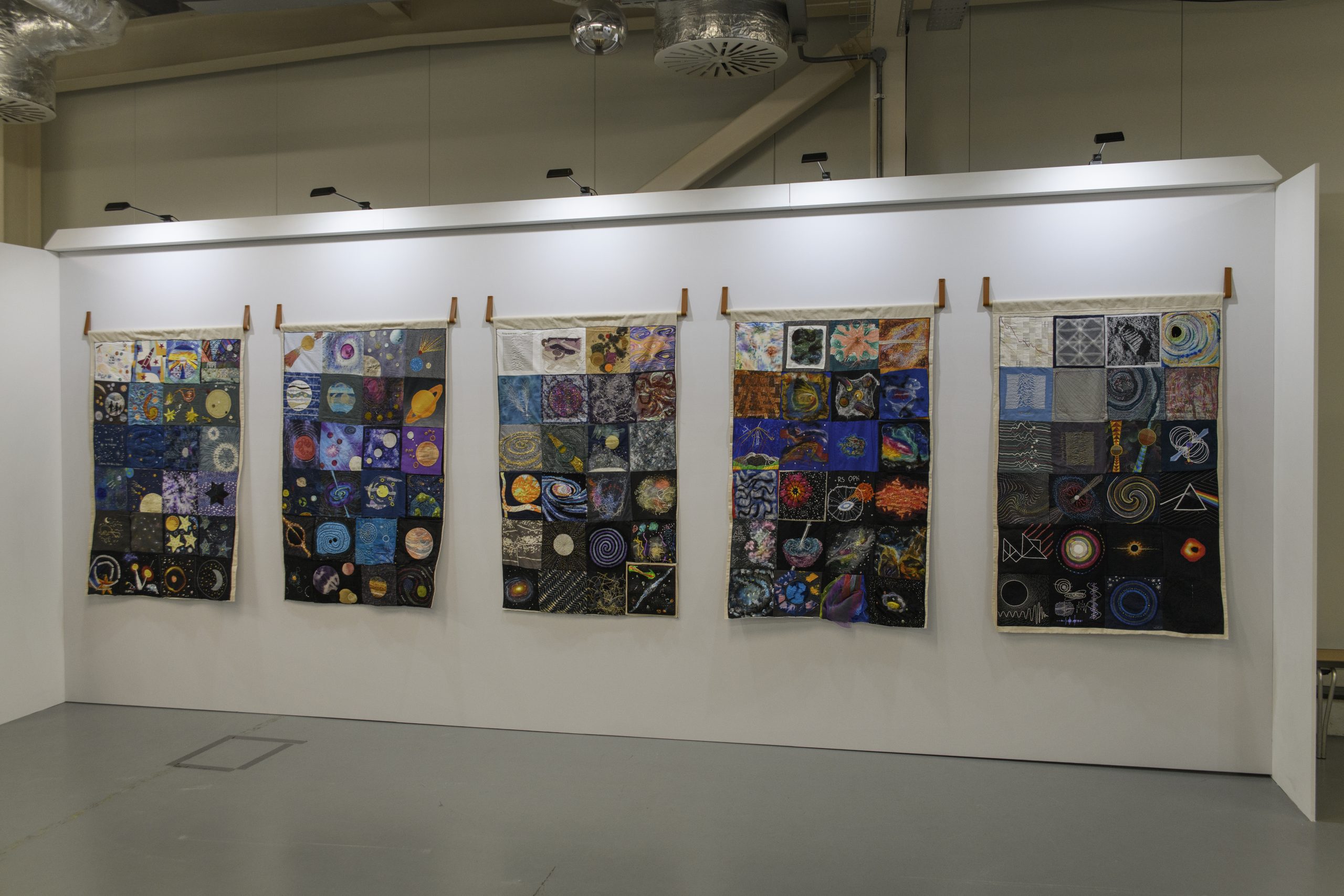 A wide shot of five of the quilts hanging side-by-side within the exhibition.