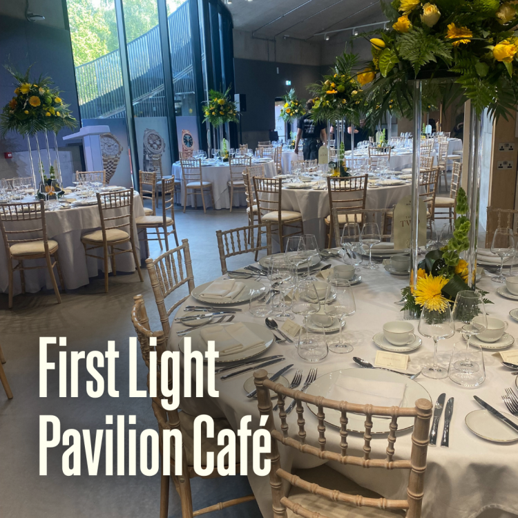 The First Light Pavilion Café set up for a cabaret-style event, with round tables laid with glassware, crockery, and floral centrepieces. The text ‘First Light Pavilion Café’ appears in the foreground of the image.
