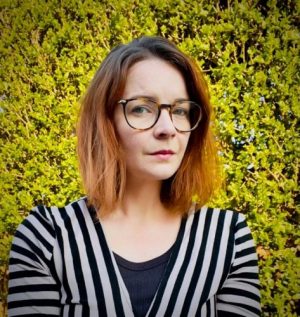 A photo of Emma Chapman, a woman wearing a black top and a black-and-white striped cardigan, standing outdoors with leafy bushes in the background.