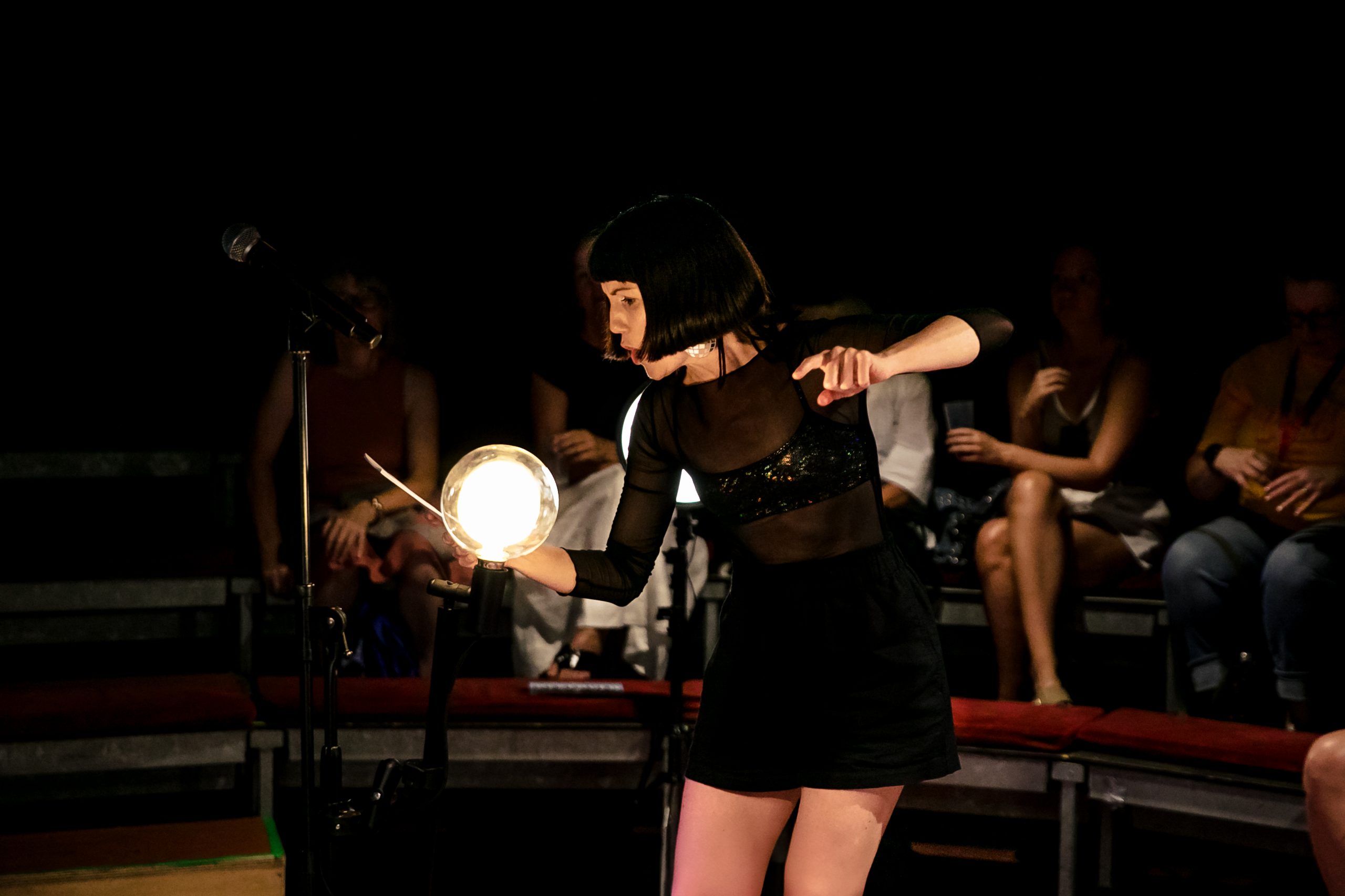 A woman in a black top and shirt reads expressively from a card on stage, illuminated by a lightbulb.