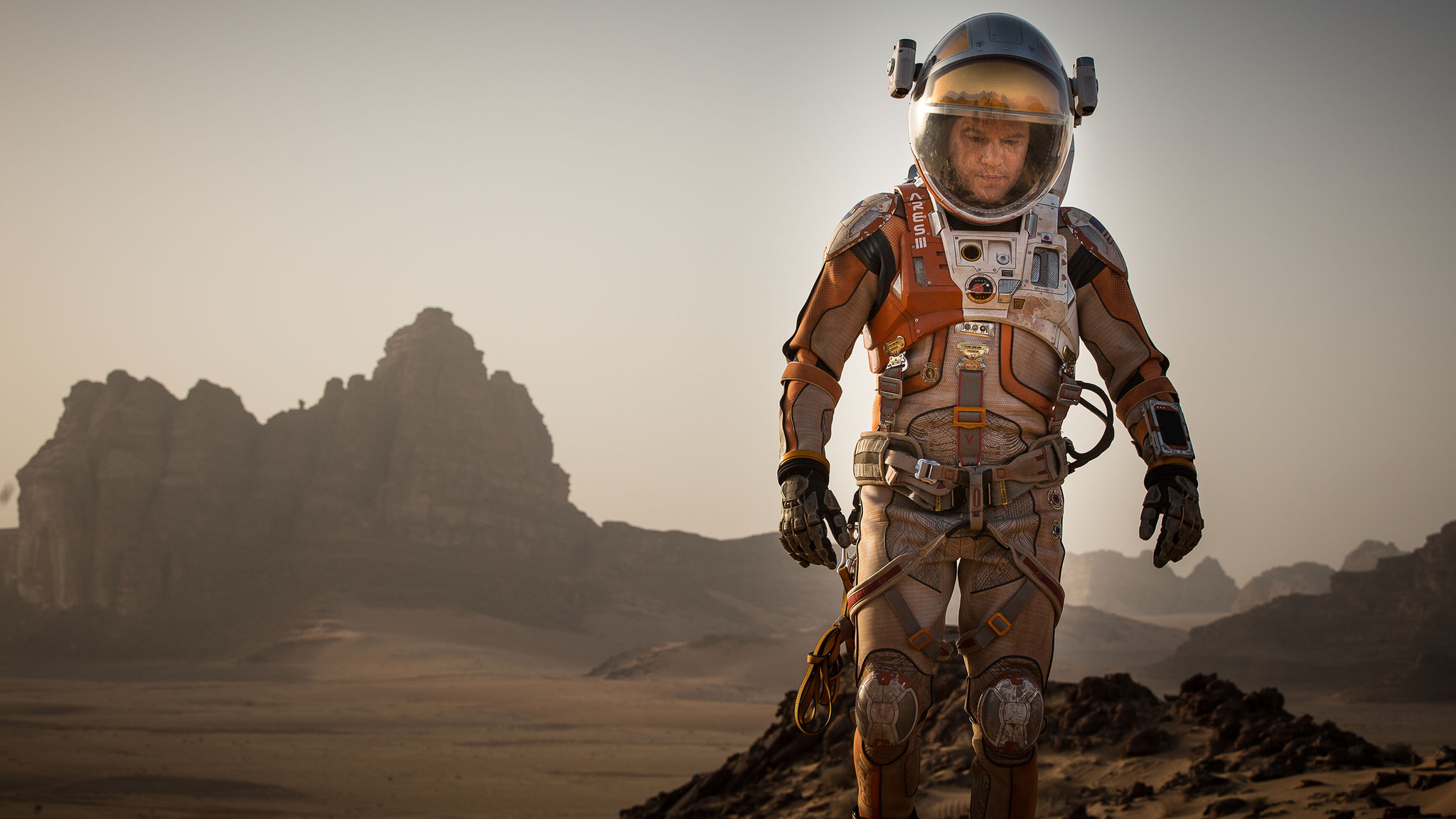 An image from the film 'The Martian', featuring Matt Damon in a red space suit, walking across the dusty, desolate landscape of Mars.