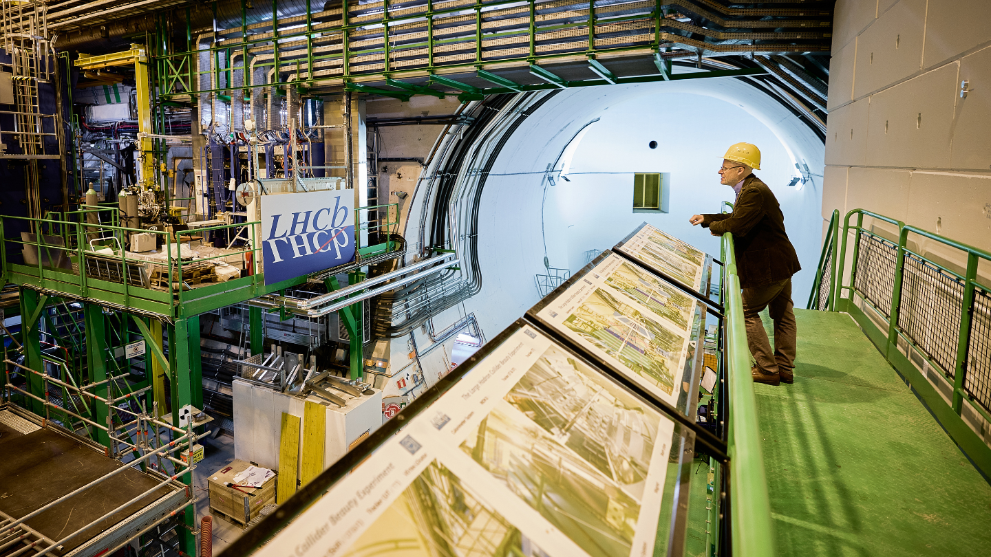 A man in a hard hat stands on a platform overlooking a large particle physics machine inside an industrial research facility.
