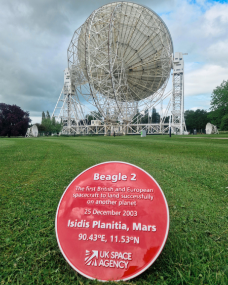 Red commemorative plaque at Jodrell Bank, displayed on grass in front of the Lovell Telescope under a cloudy sky.