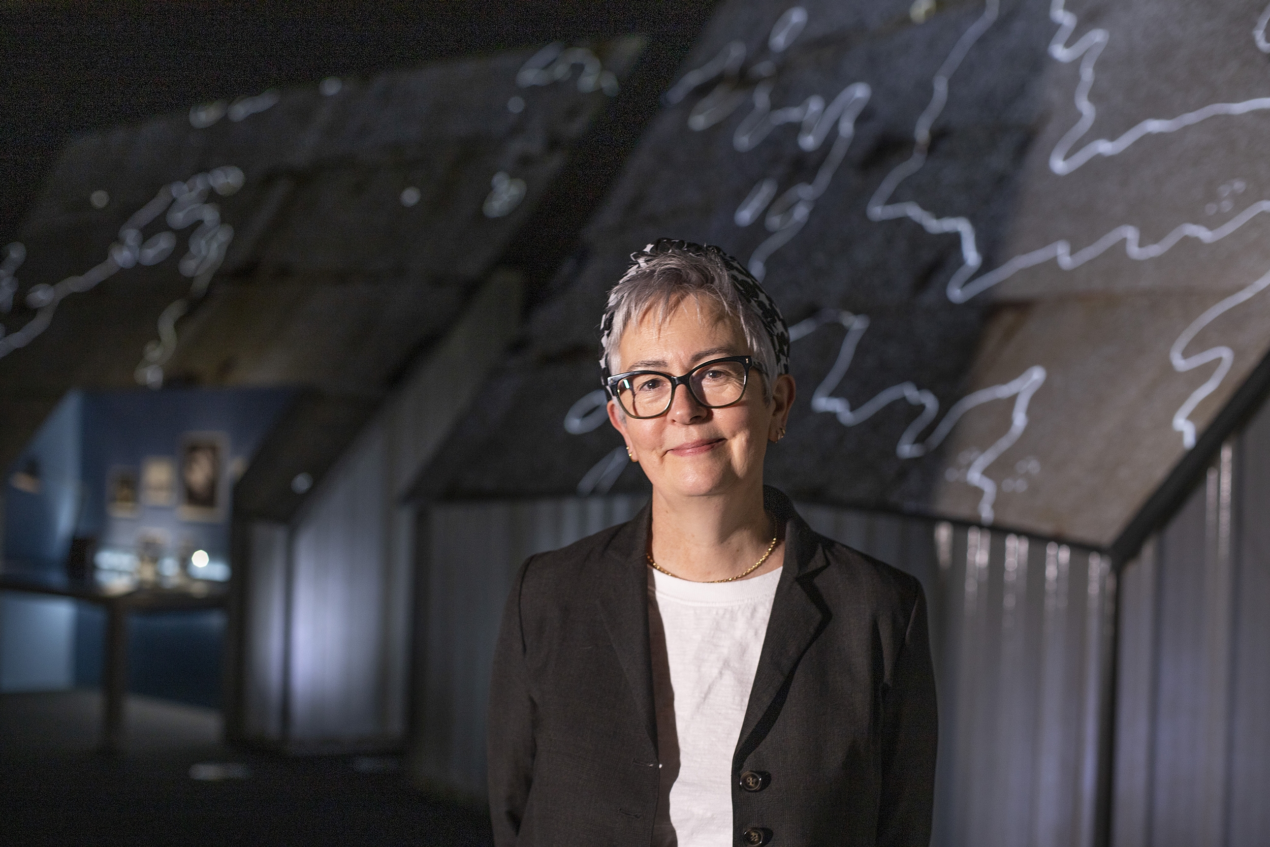 Teresa Anderson, a woman with short grey hair and glasses, stands in the First Light Pavilion exhibition.